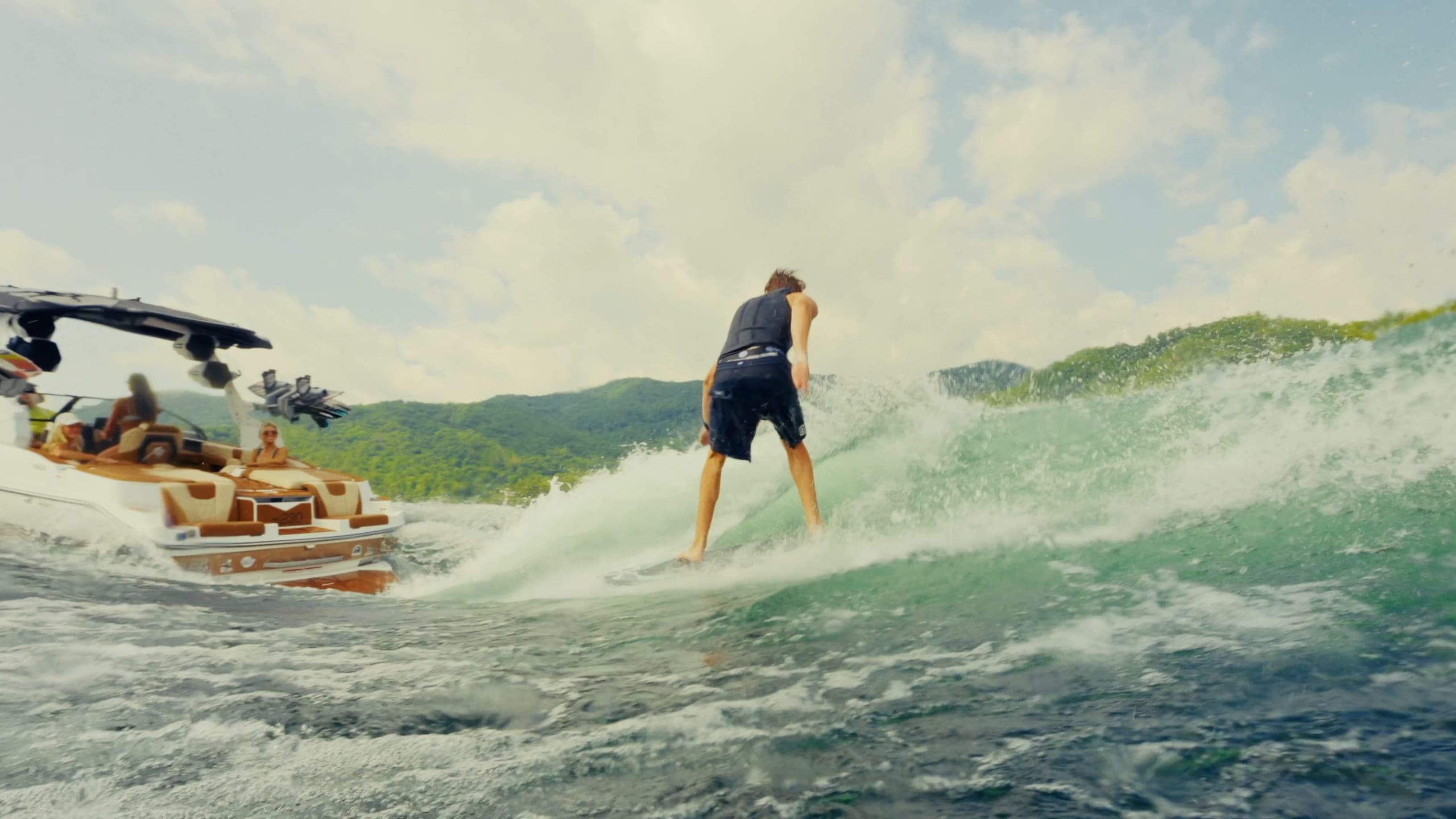 A rider surfs the wave behind a Malibu M Series boat, then the boat rests on calm water in the late sun