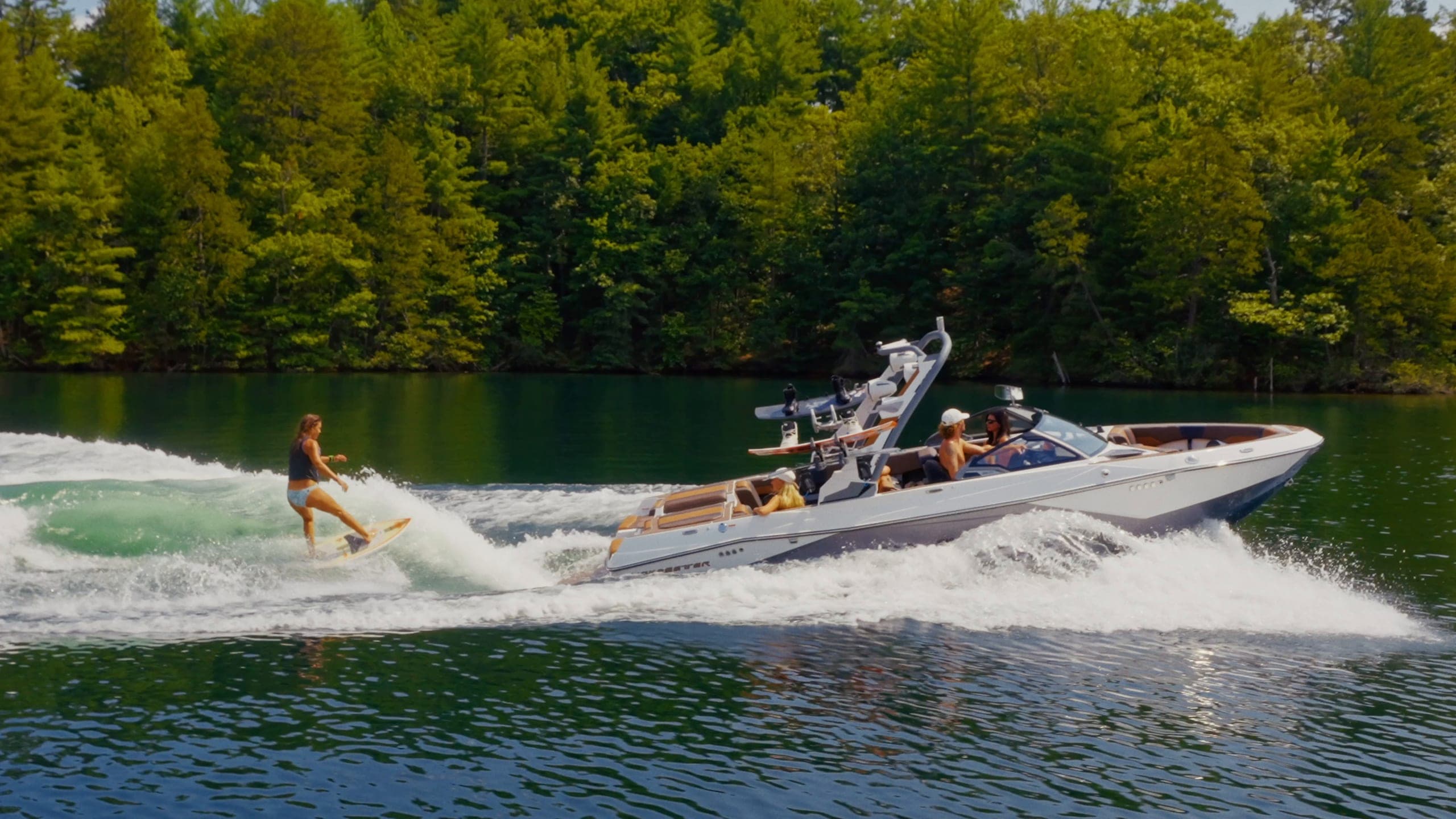 A Malibu 23 LSV at speed at sunset, a swimmer beside it, a rider surfing behind it, and the boat turning seen from above