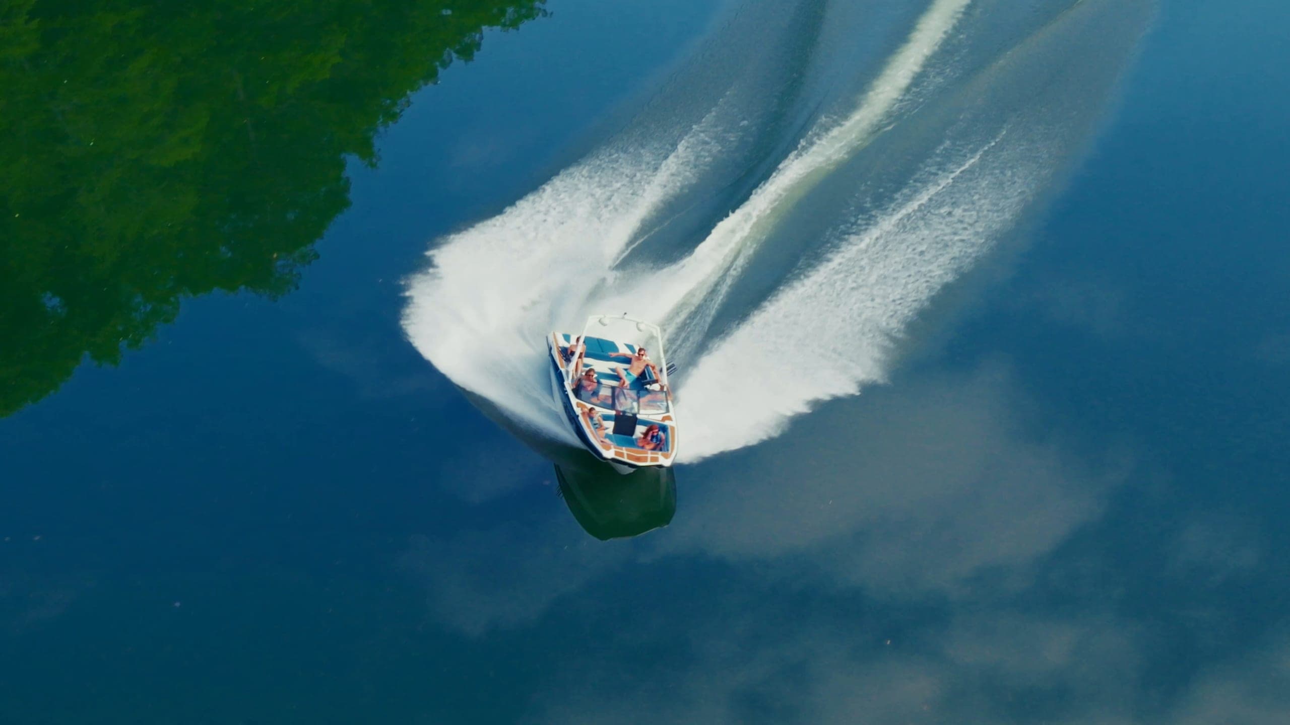 Friends aboard an Axis A225 watch a rider surf, a wakeboarder rides, and the boat carves a turn seen from above
