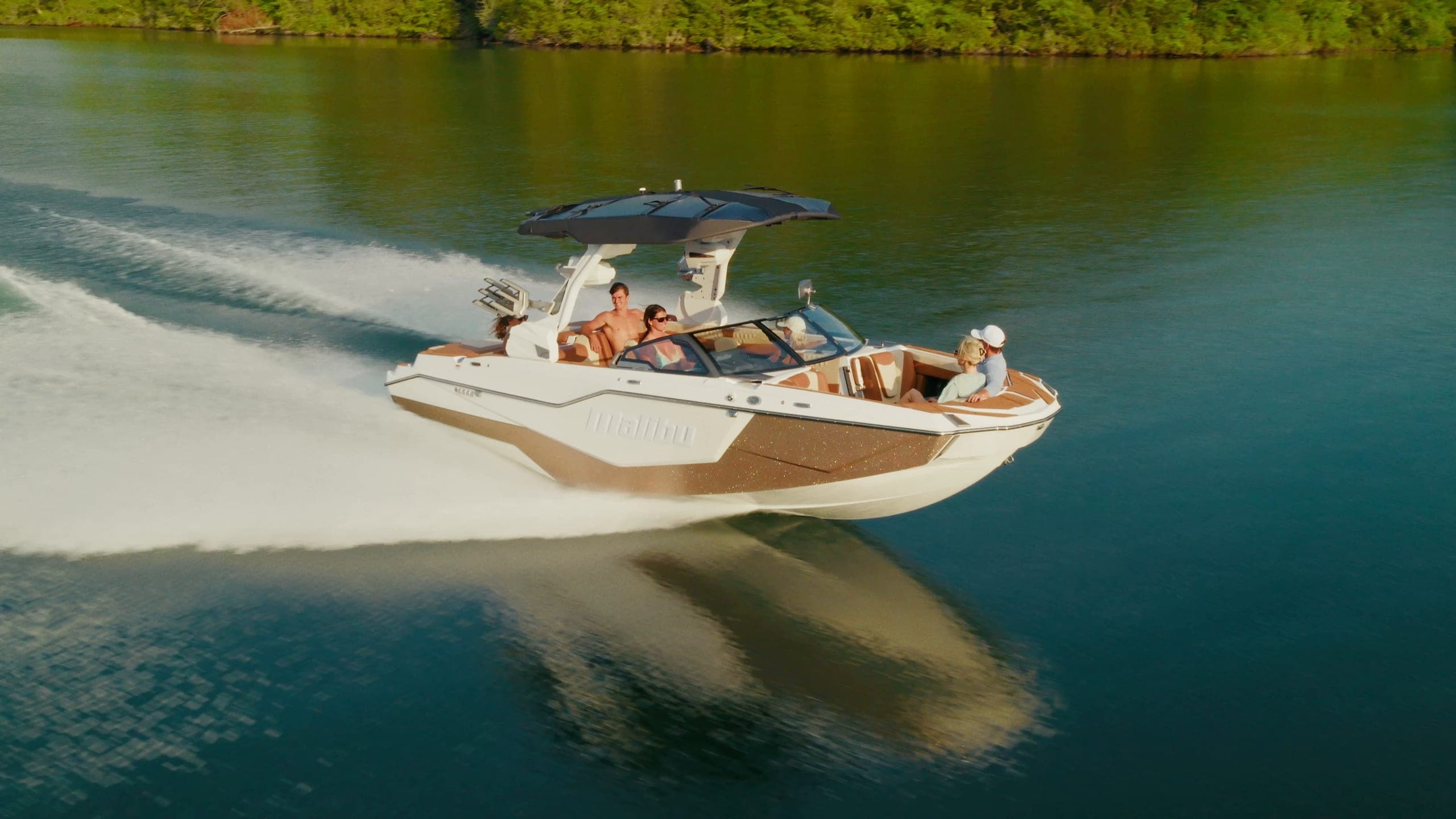A Malibu M230 at speed on calm water, the driver at the touchscreen helm, and a rider in the air off its wave